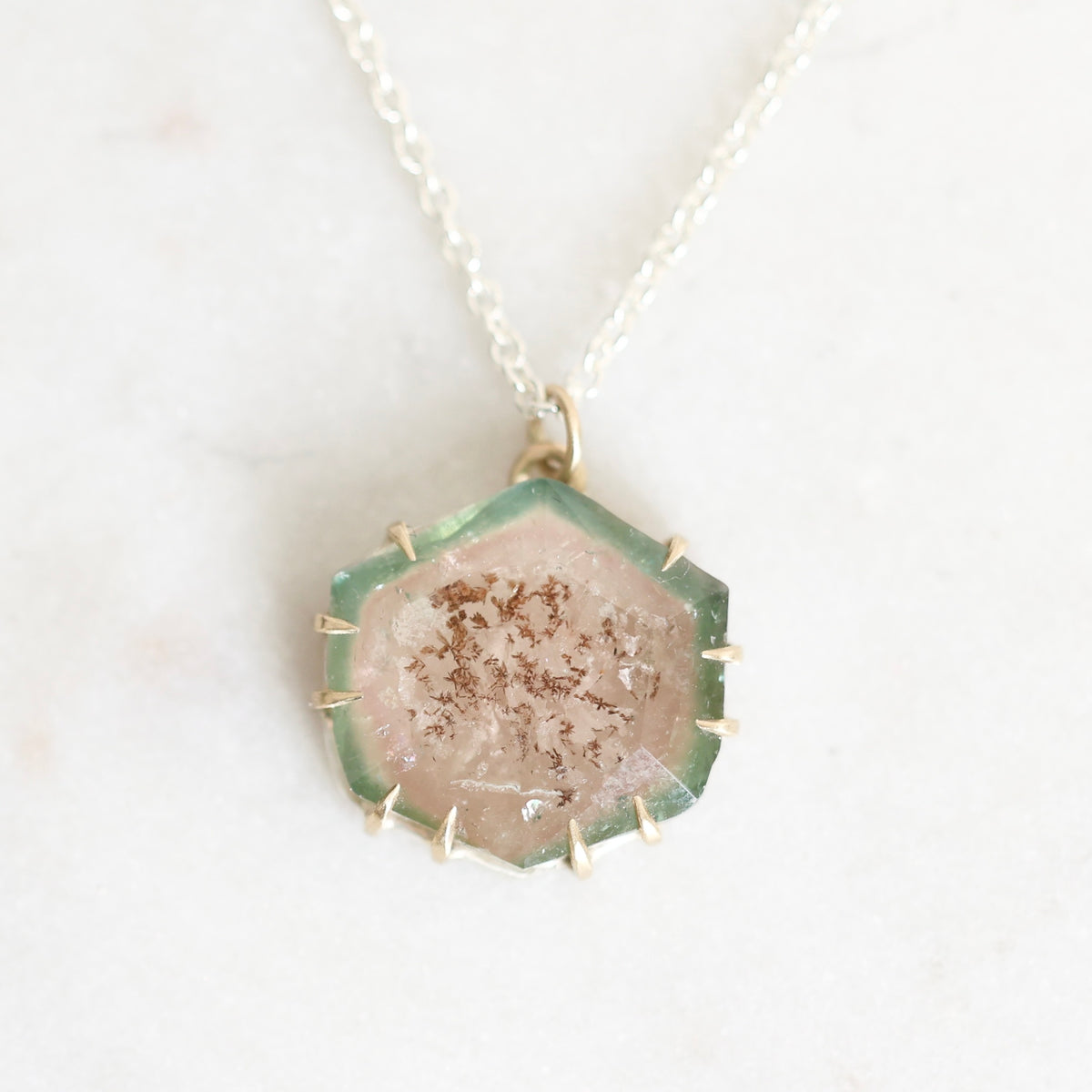 A hexagonal druzy crystal pendant necklace on a delicate silver chain, displayed on a light marble surface. The pendant features a pale pinkish-beige crystalline center with brown mineral inclusions, surrounded by a green-tinted glassy border and a gold-toned bezel.