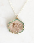 A hexagonal druzy crystal pendant necklace on a delicate silver chain, displayed on a light marble surface. The pendant features a pale pinkish-beige crystalline center with brown mineral inclusions, surrounded by a green-tinted glassy border and a gold-toned bezel.