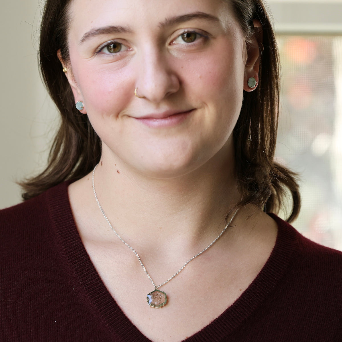 A woman with brown hair wearing a silver necklace with a circular pendant and matching stud earrings, photographed in a close-up portrait.