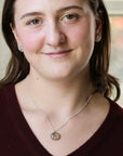 A woman with brown hair wearing a silver necklace with a circular pendant and matching stud earrings, photographed in a close-up portrait.