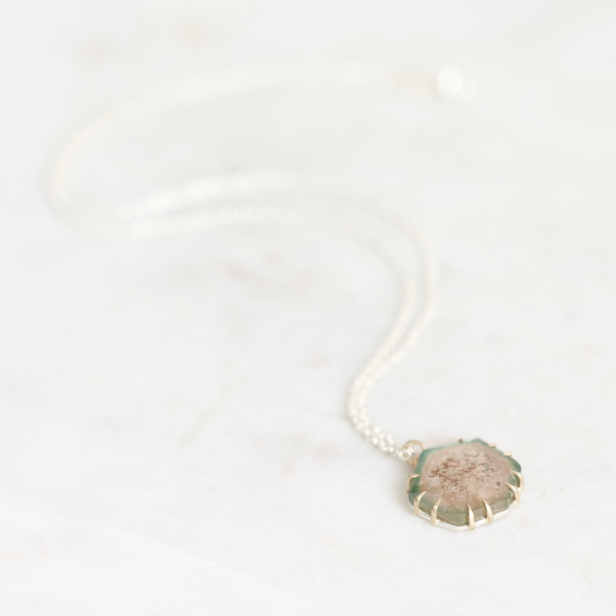 A delicate silver chain necklace with a small, scalloped circular pendant featuring a textured, frosted stone in muted green, pink, and beige tones, resting on a light marble surface.