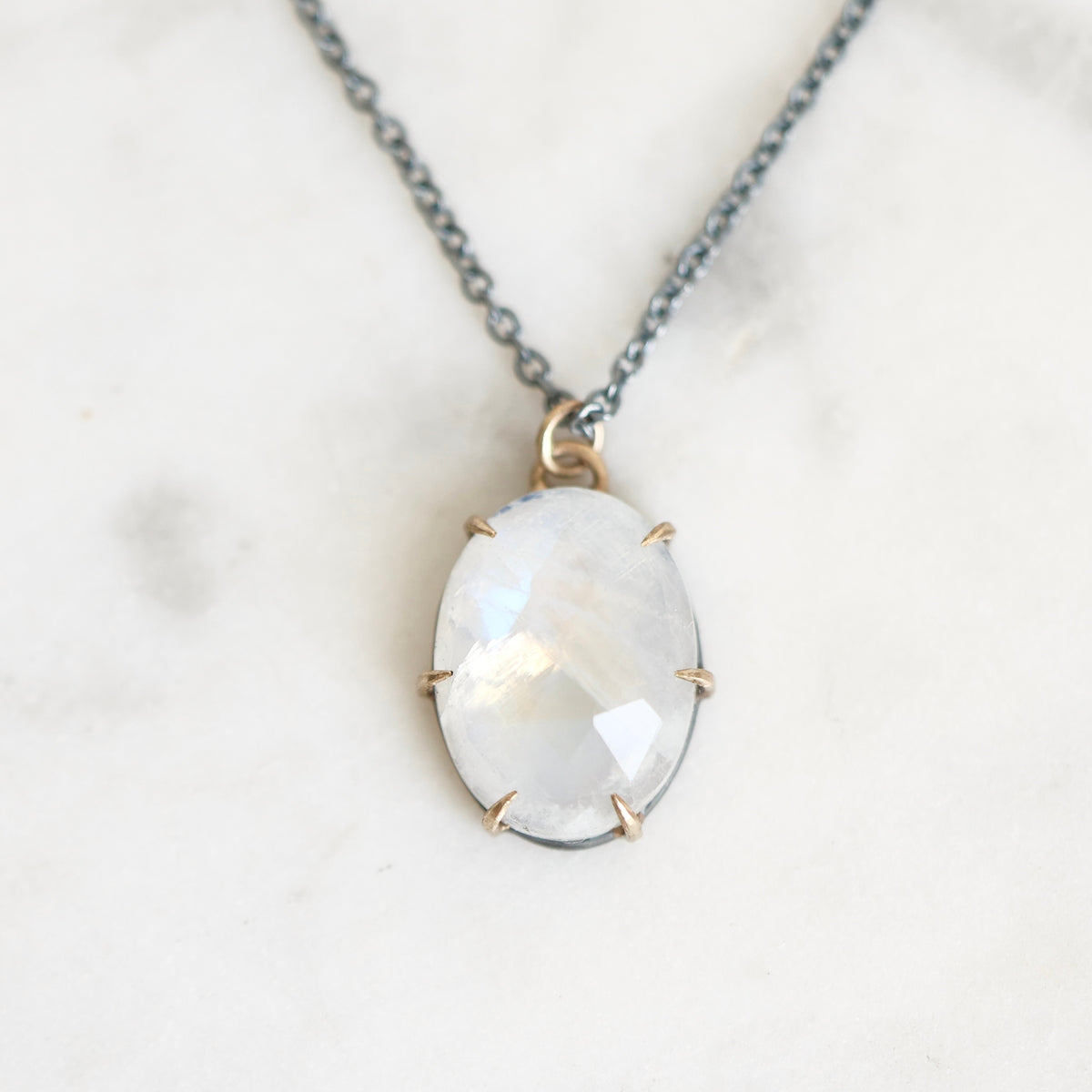 An oval pendant necklace featuring a faceted white moonstone with subtle iridescent sheen, set in a gold prong setting on a dark gray chain, displayed on a light marble surface.