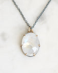 An oval pendant necklace featuring a faceted white moonstone with subtle iridescent sheen, set in a gold prong setting on a dark gray chain, displayed on a light marble surface.