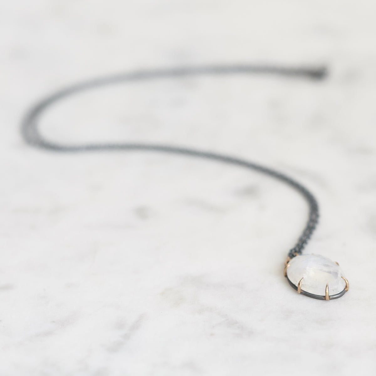 A delicate dark gray chain necklace with a small oval pendant featuring a milky white cabochon stone in a gold-toned bezel, resting on a white marble surface.