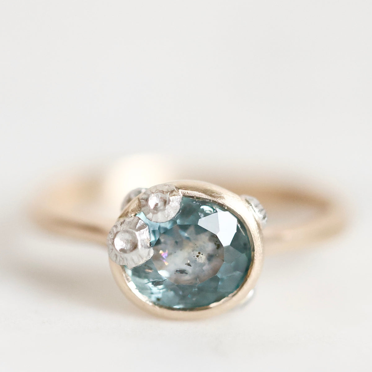 A close-up shot of a gold ring featuring a round faceted light blue gemstone, accented by small clear stones on the shoulders, set against a soft, out-of-focus light gray background.