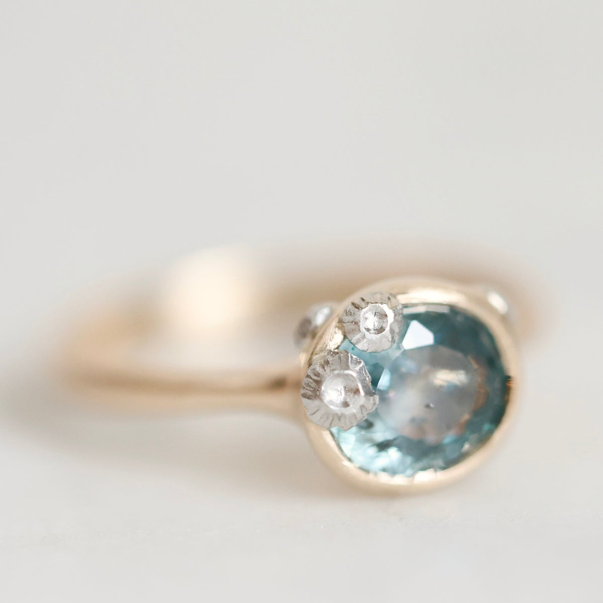 A close-up shot of a gold ring featuring a large oval light blue gemstone and three small clear accent stones, set against a soft, light gray background.