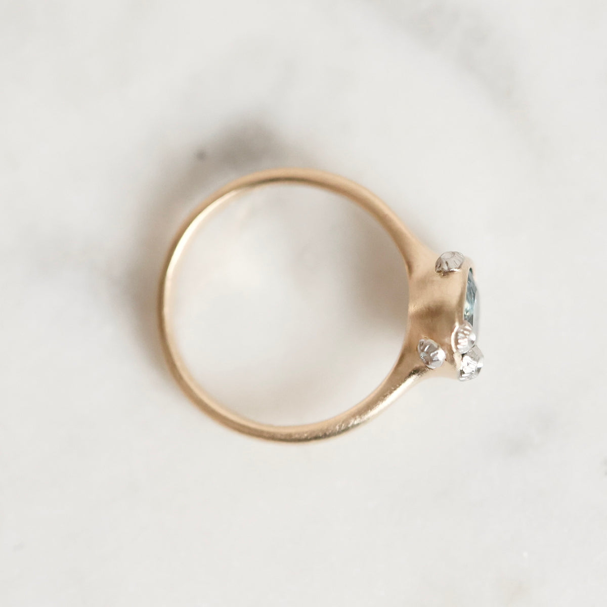 A gold ring with a small cluster of clear gemstones on the side, photographed from above on a light marble surface.