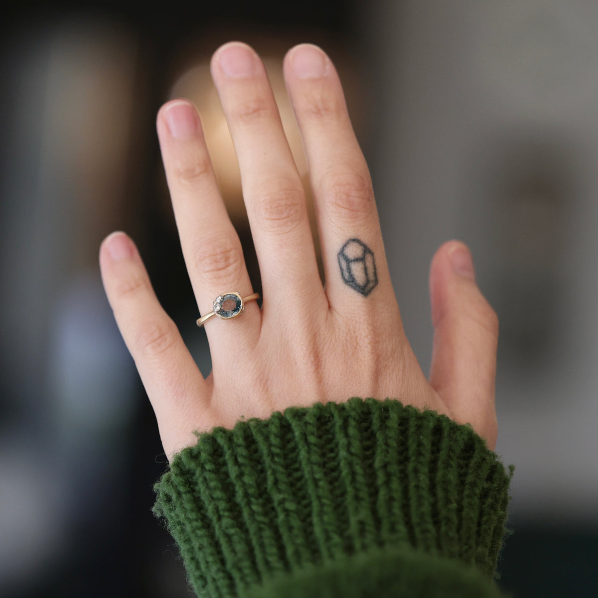 A close-up of a person's hand wearing a silver ring with a round blue gemstone, set against a blurred background with a green knit sweater cuff visible.