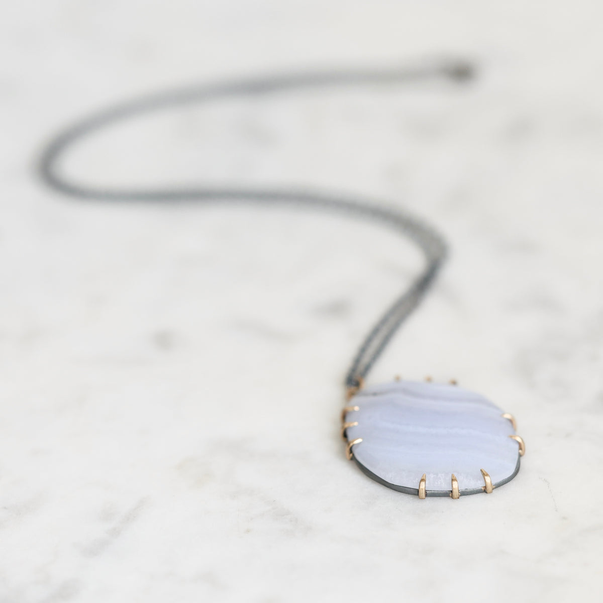 A delicate necklace featuring a light blue-gray agate stone pendant with gold prong settings and a dark gray cord, resting on a white marble surface.