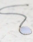 A delicate necklace featuring a light blue-gray agate stone pendant with gold prong settings and a dark gray cord, resting on a white marble surface.