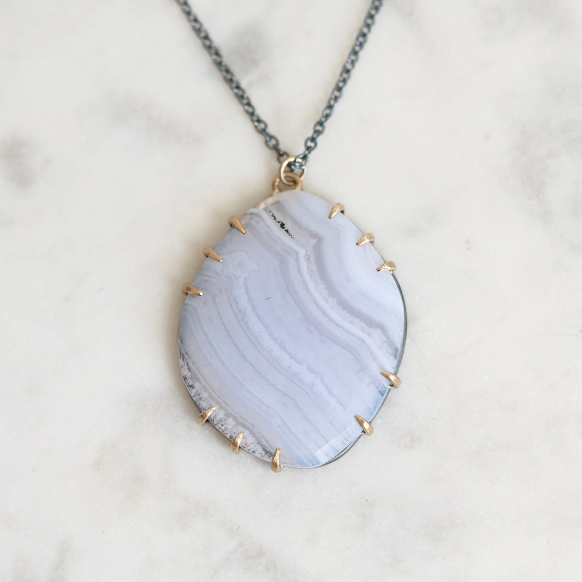A delicate necklace featuring a light blue-gray agate stone pendant with gold prong settings and a dark gray cord, resting on a white marble surface.
