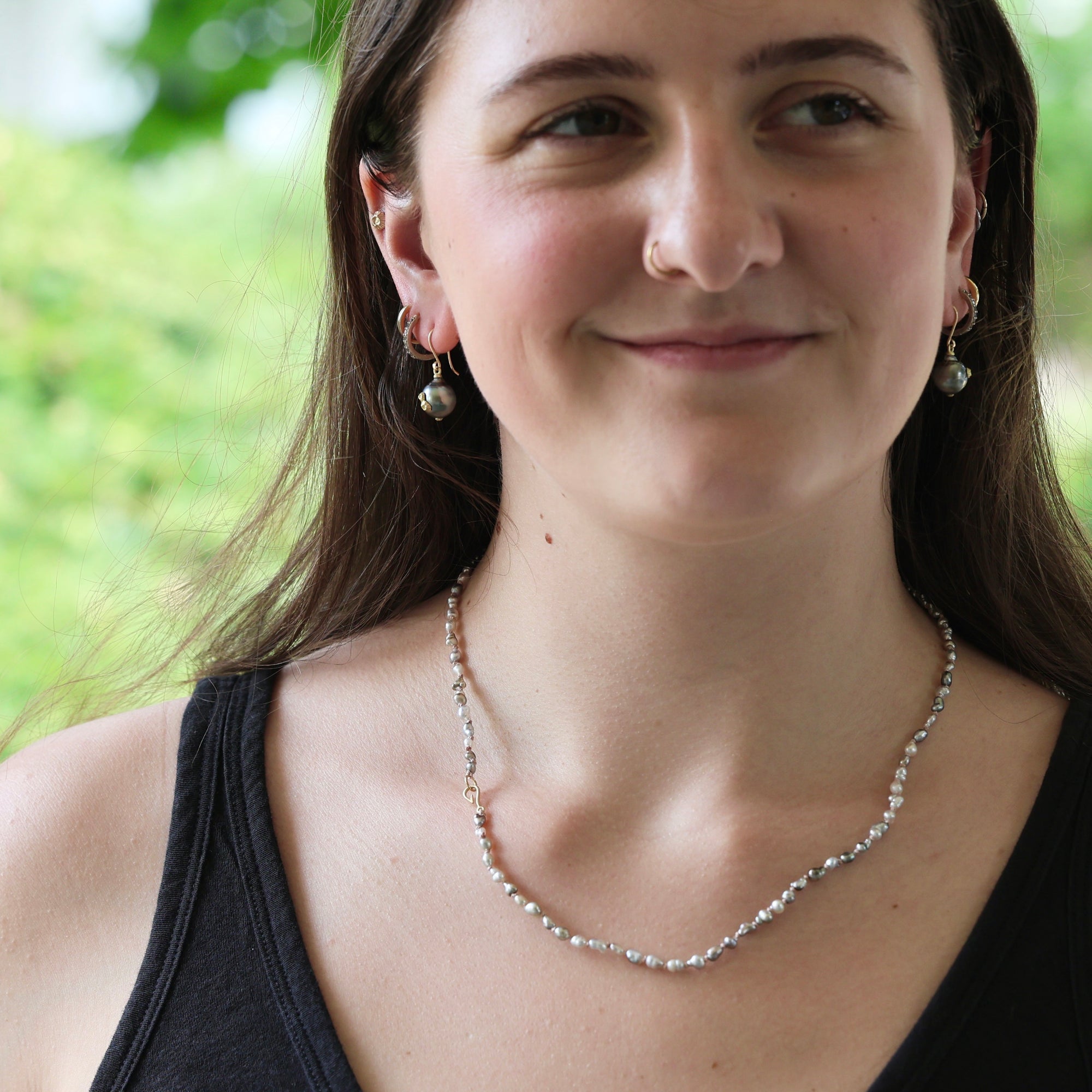 A woman wearing a delicate silver beaded necklace and matching silver drop earrings, photographed outdoors with a soft green blurred background.
