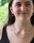 A woman wearing a delicate silver beaded necklace and matching silver drop earrings, photographed outdoors with a soft green blurred background.