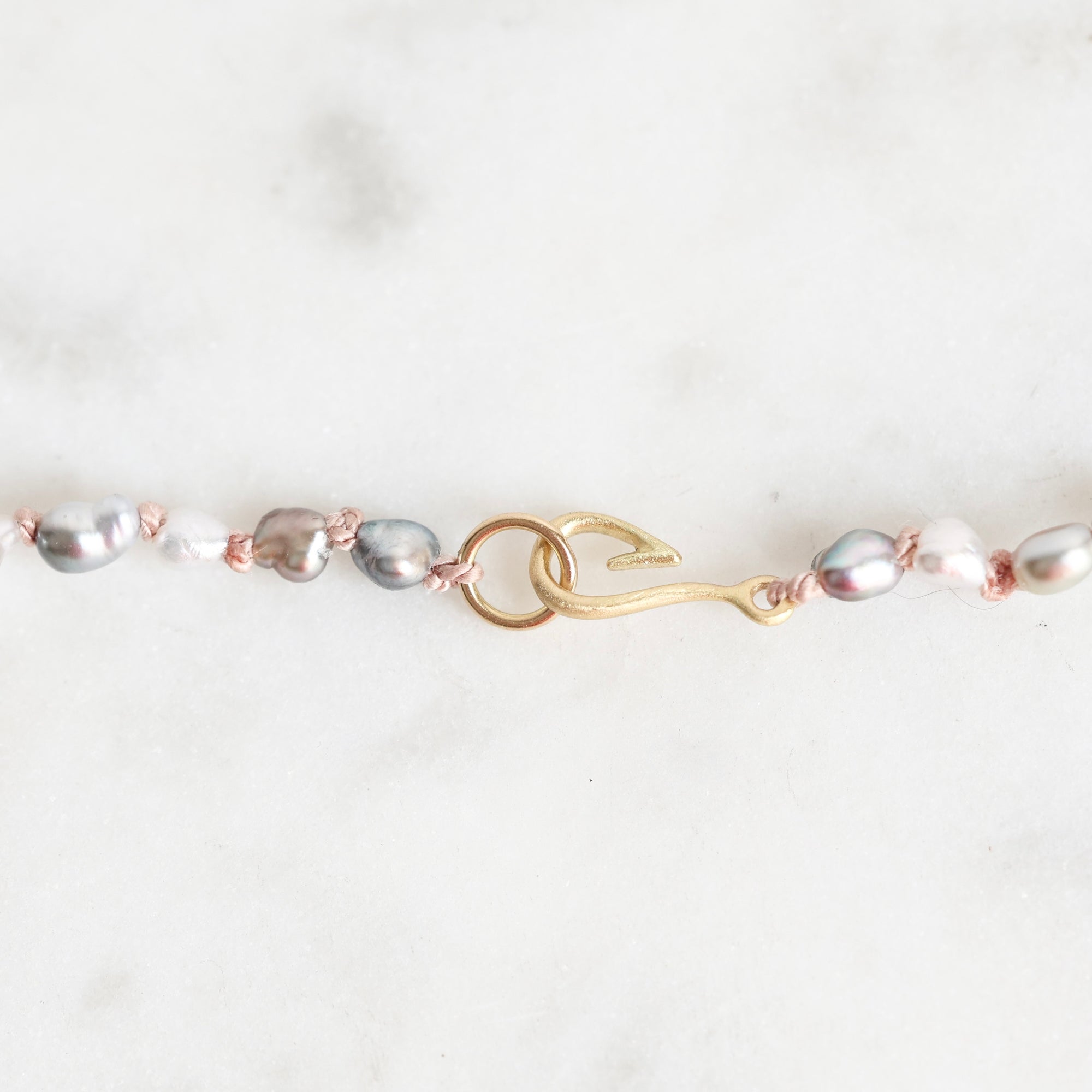 A close-up of a delicate jewelry piece featuring baroque pearls in silver, white, and gray tones, accented with rose gold wire and a gold hook-shaped charm, set against a light marble background.
