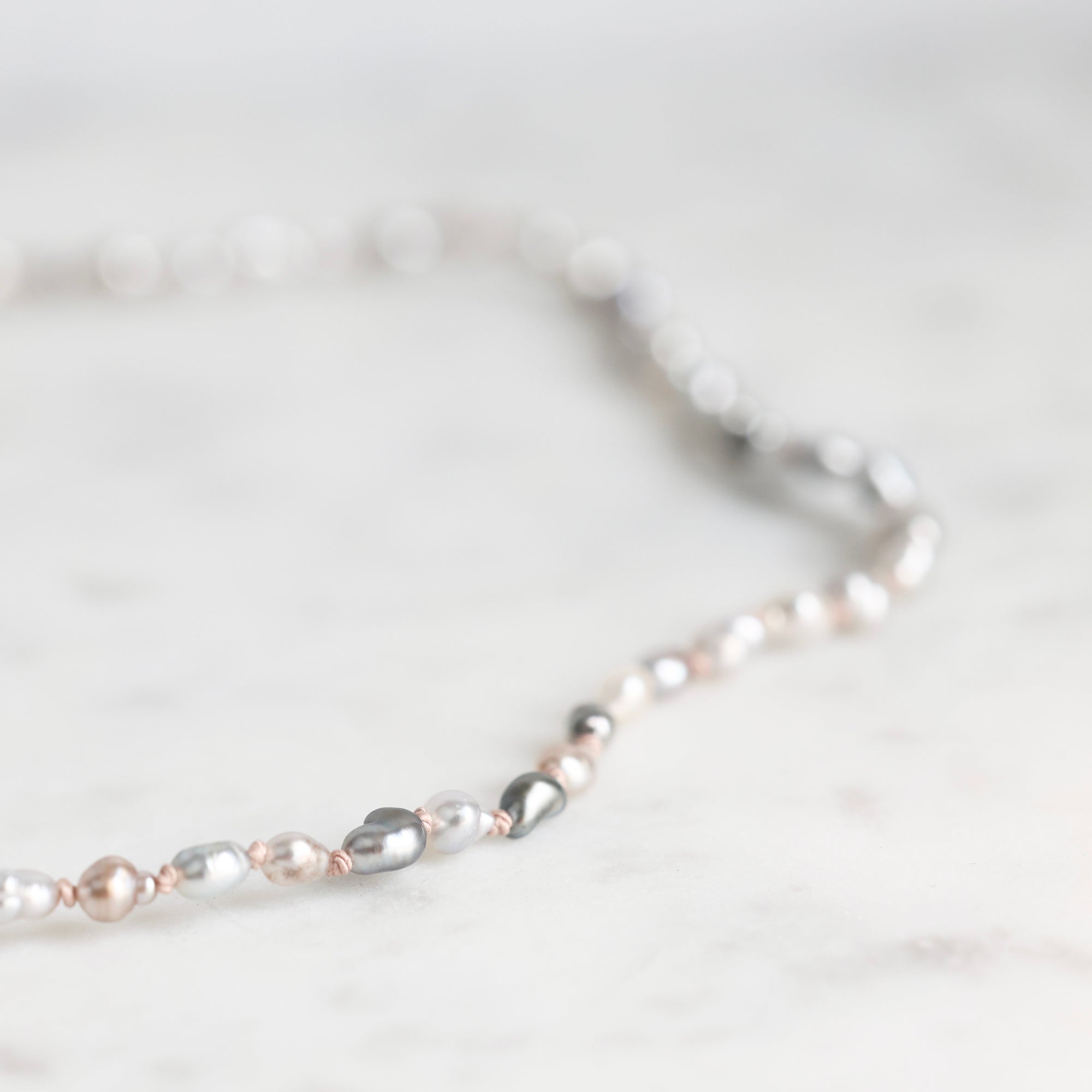 A close-up of a beaded strand necklace with mixed silver, white, and pale pink pearls on a light marble surface.
