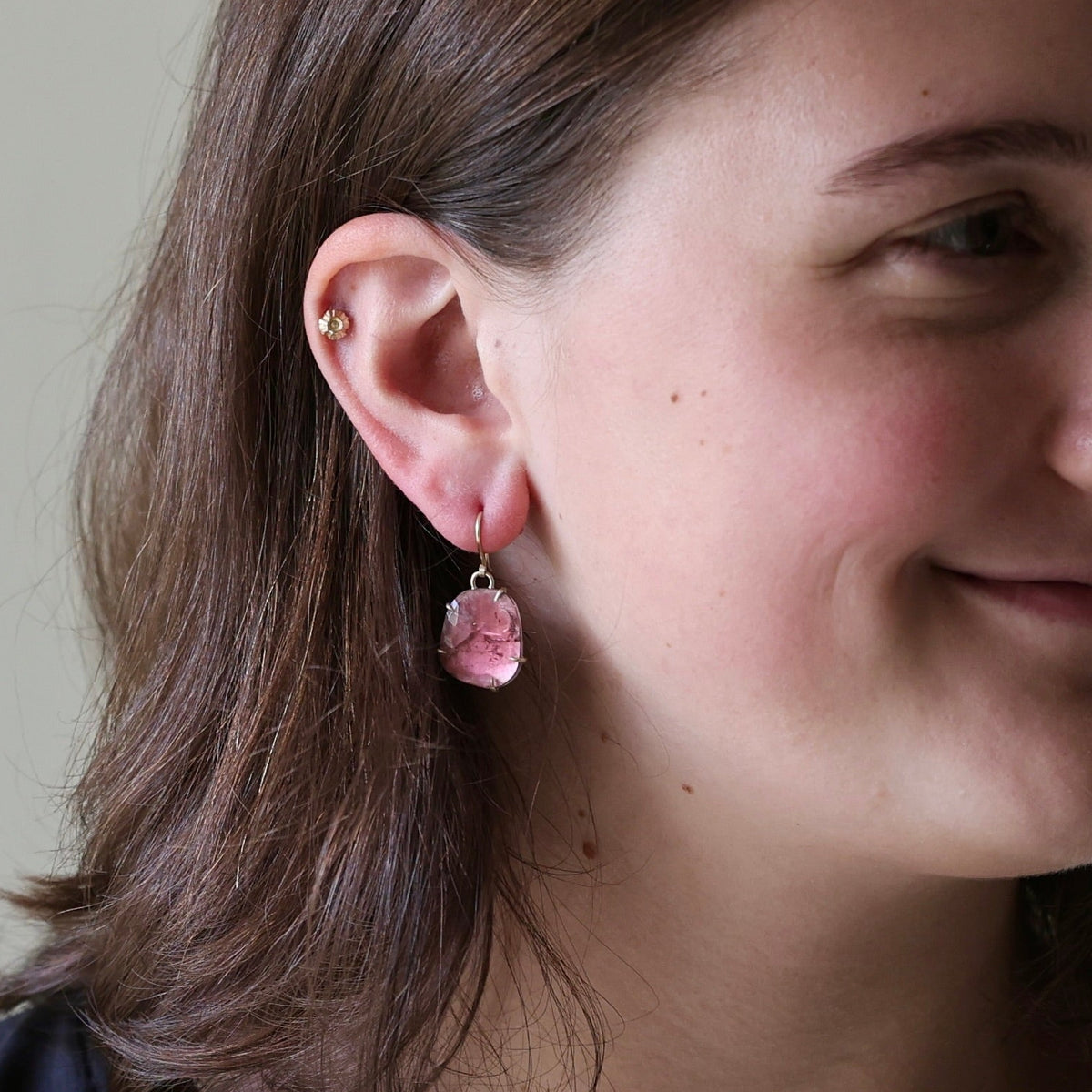 A woman with brown hair wearing a small pink dangling earring, shown in a close-up side profile against a soft neutral indoor background.