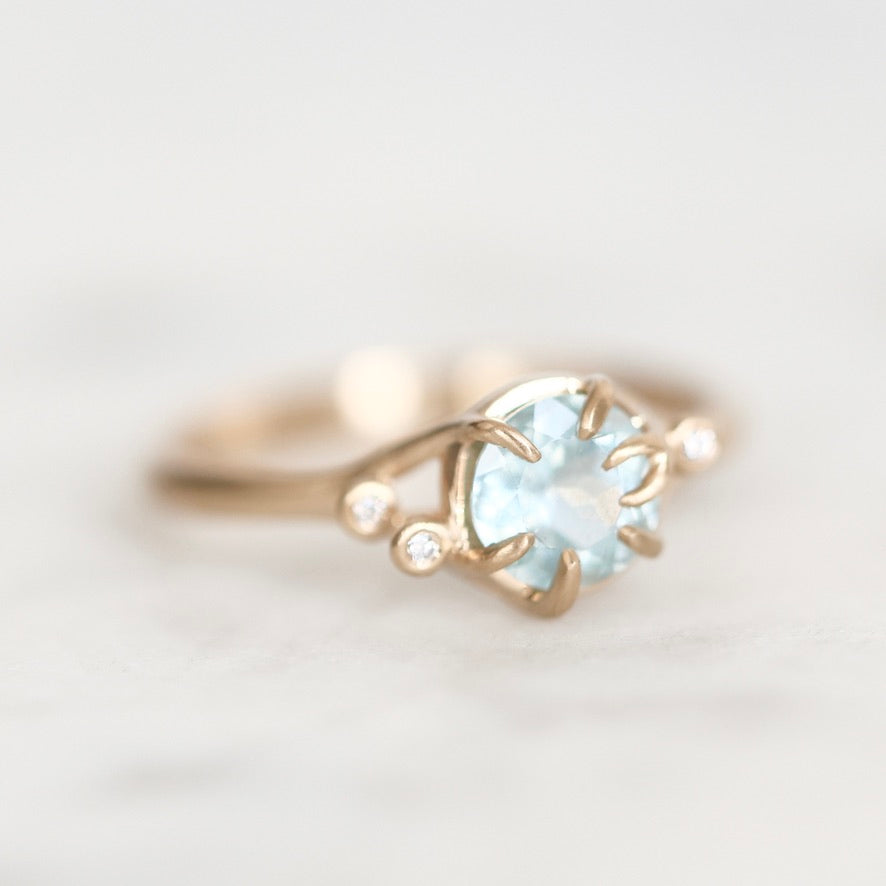 A delicate gold ring featuring a round light blue gemstone in a prong setting, accented by two small clear stones on the band, photographed on a soft white background.