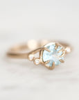 A delicate gold ring featuring a round light blue gemstone in a prong setting, accented by two small clear stones on the band, photographed on a soft white background.