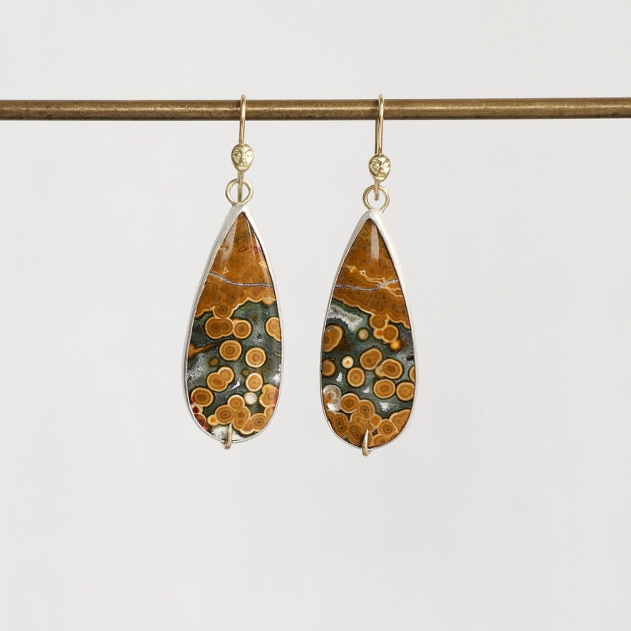 A pair of teardrop-shaped drop earrings with polished brown and orange stone featuring circular patterns, set in silver with gold hooks, hanging from a horizontal gold rod against a light gray background.