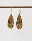 A pair of teardrop-shaped drop earrings with polished brown and orange stone featuring circular patterns, set in silver with gold hooks, hanging from a horizontal gold rod against a light gray background.