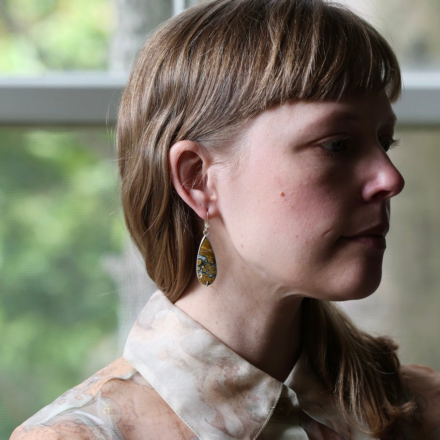 A woman in profile wearing a dangling teardrop earring with a marbled yellow, blue, and green stone in a gold-toned setting, photographed indoors near a window.