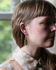 A woman in profile wearing a dangling teardrop earring with a marbled yellow, blue, and green stone in a gold-toned setting, photographed indoors near a window.