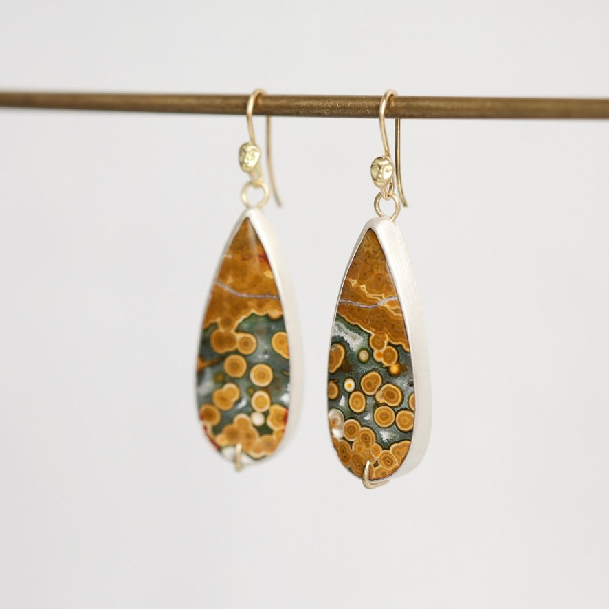 Teardrop-shaped dangle earrings with a natural orange and green agate pattern, set in silver and suspended from gold hooks, hanging from a wooden rod against a light gray background.