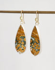 Teardrop-shaped dangle earrings with a natural orange and green agate pattern, set in silver and suspended from gold hooks, hanging from a wooden rod against a light gray background.