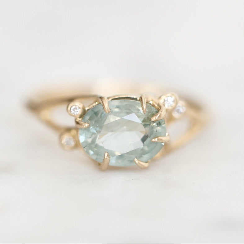 A yellow gold ring featuring a large oval-cut light blue gemstone in a prong setting, accented by two small round clear stones on the band, photographed on a soft white background.