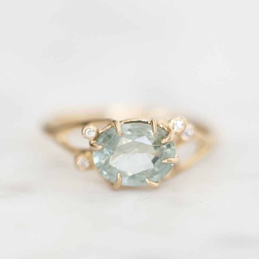A yellow gold ring featuring a large oval-cut light blue gemstone in a prong setting, accented by two small round clear stones on the band, photographed on a soft white background.