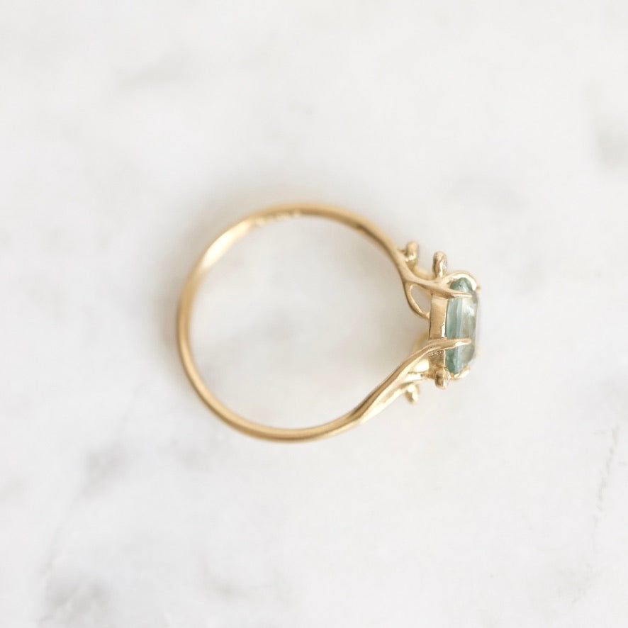 A gold ring with a small rectangular light green gemstone, photographed from above on a white marble surface.