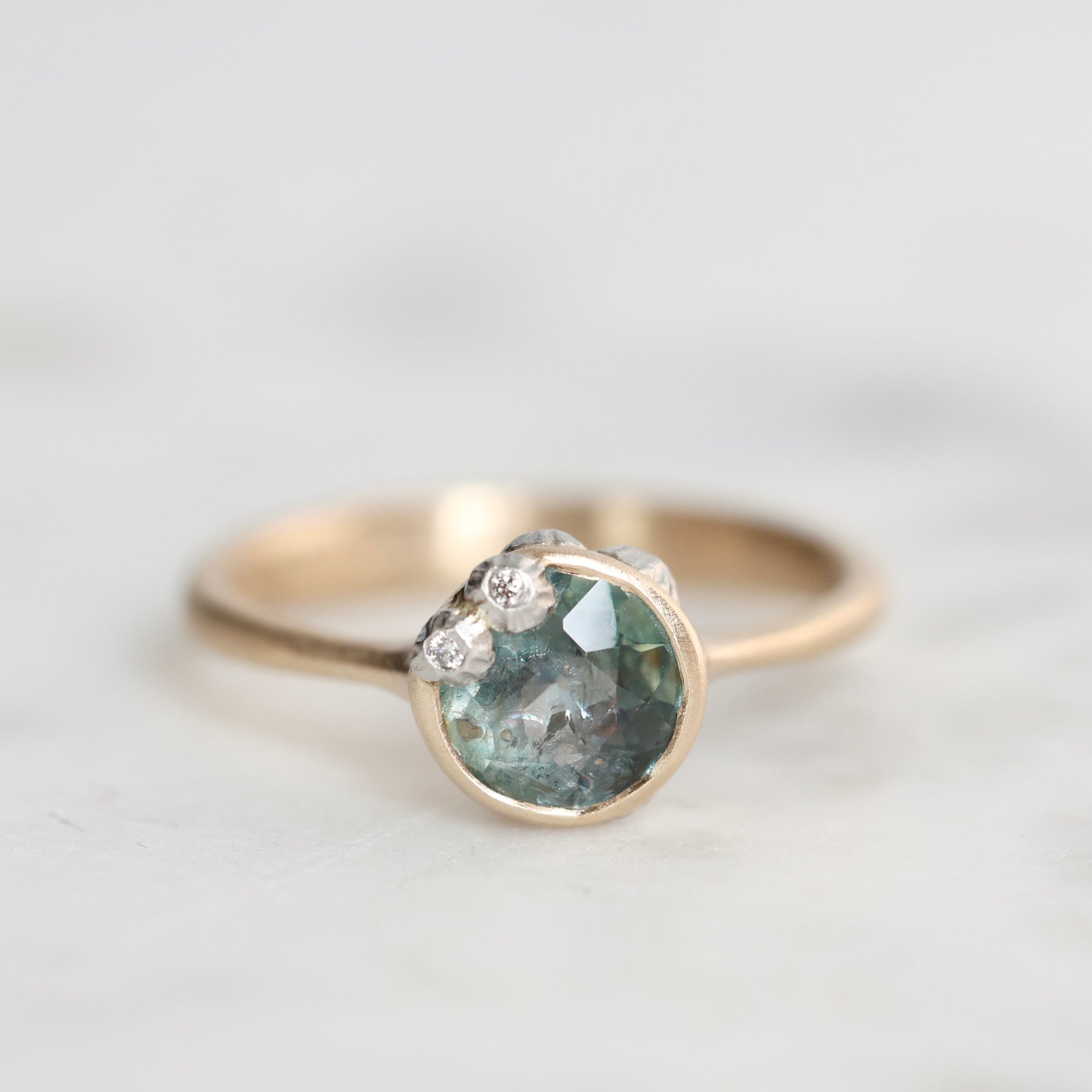 A gold ring featuring a round, faceted light blue-green gemstone with small barnacless on the upper left, set against a soft, light gray background.