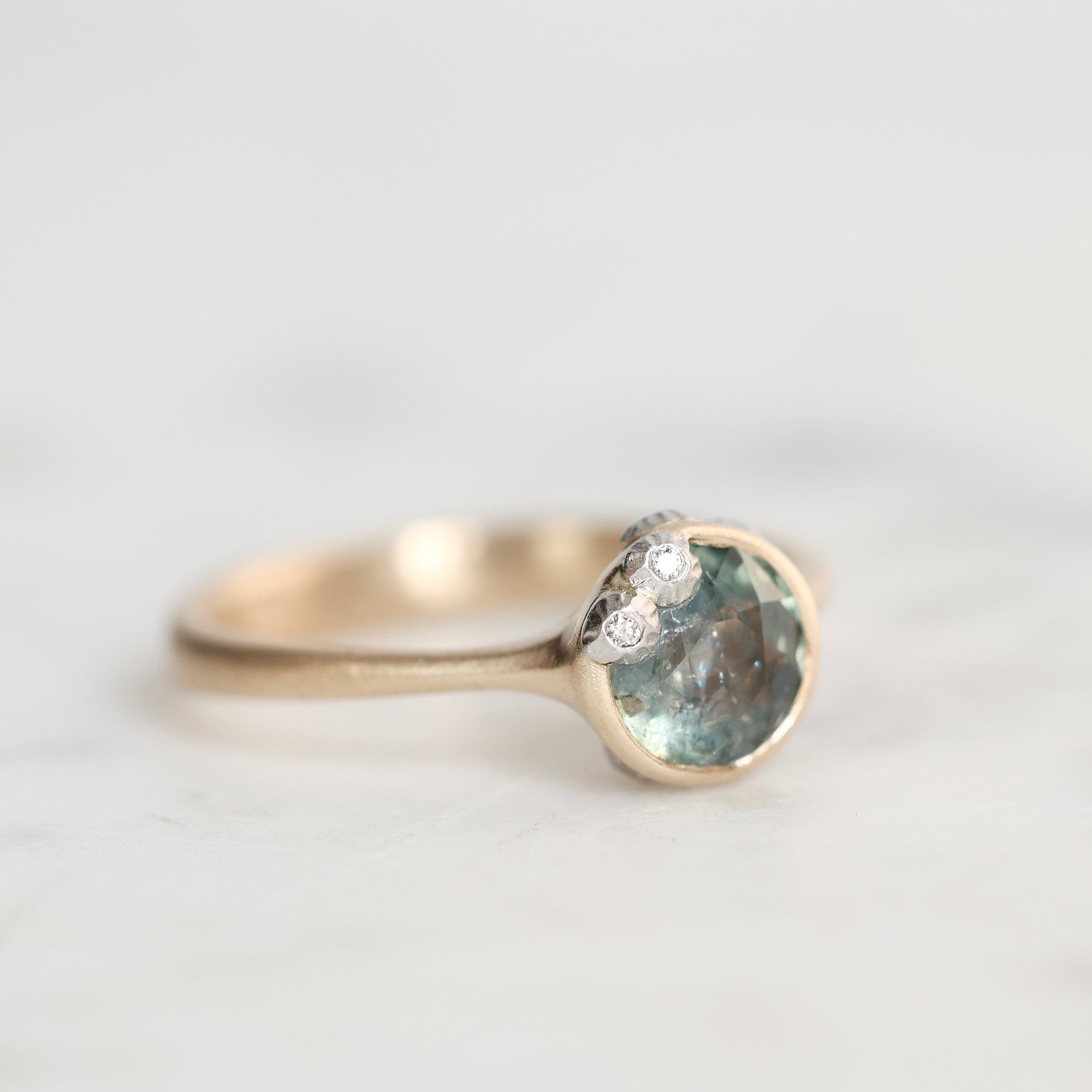 A close-up shot of a gold ring featuring a round light blue gemstone and a small cluster of barnacles on the band, set against a soft, light gray background.