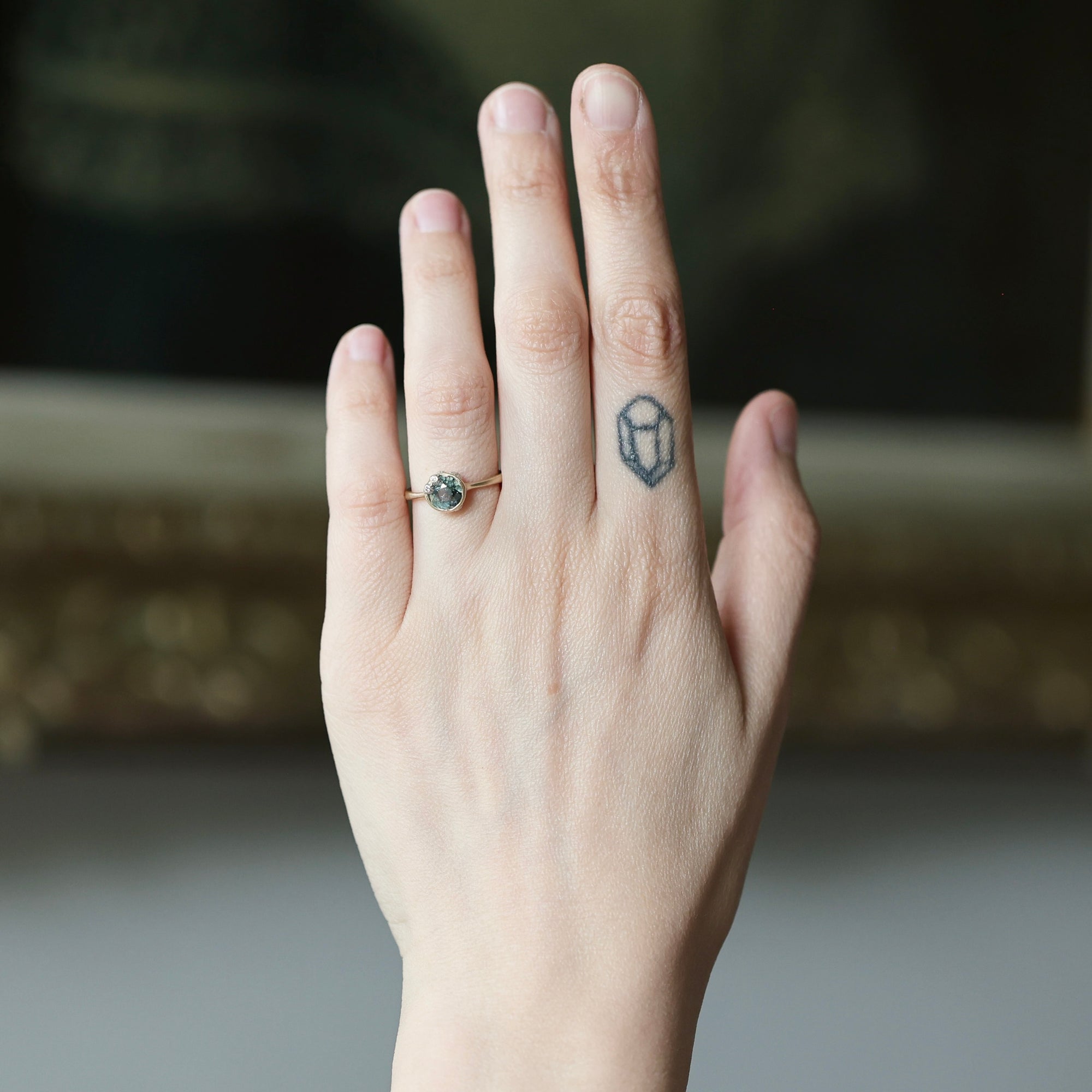A close-up of a person's hand showing a delicate gold ring with a small round dark gemstone on the ring finger, with a small geometric tattoo on the middle finger. The background is a soft, out-of-focus dark interior.