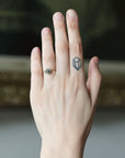 A close-up of a person's hand showing a delicate gold ring with a small round dark gemstone on the ring finger, with a small geometric tattoo on the middle finger. The background is a soft, out-of-focus dark interior.
