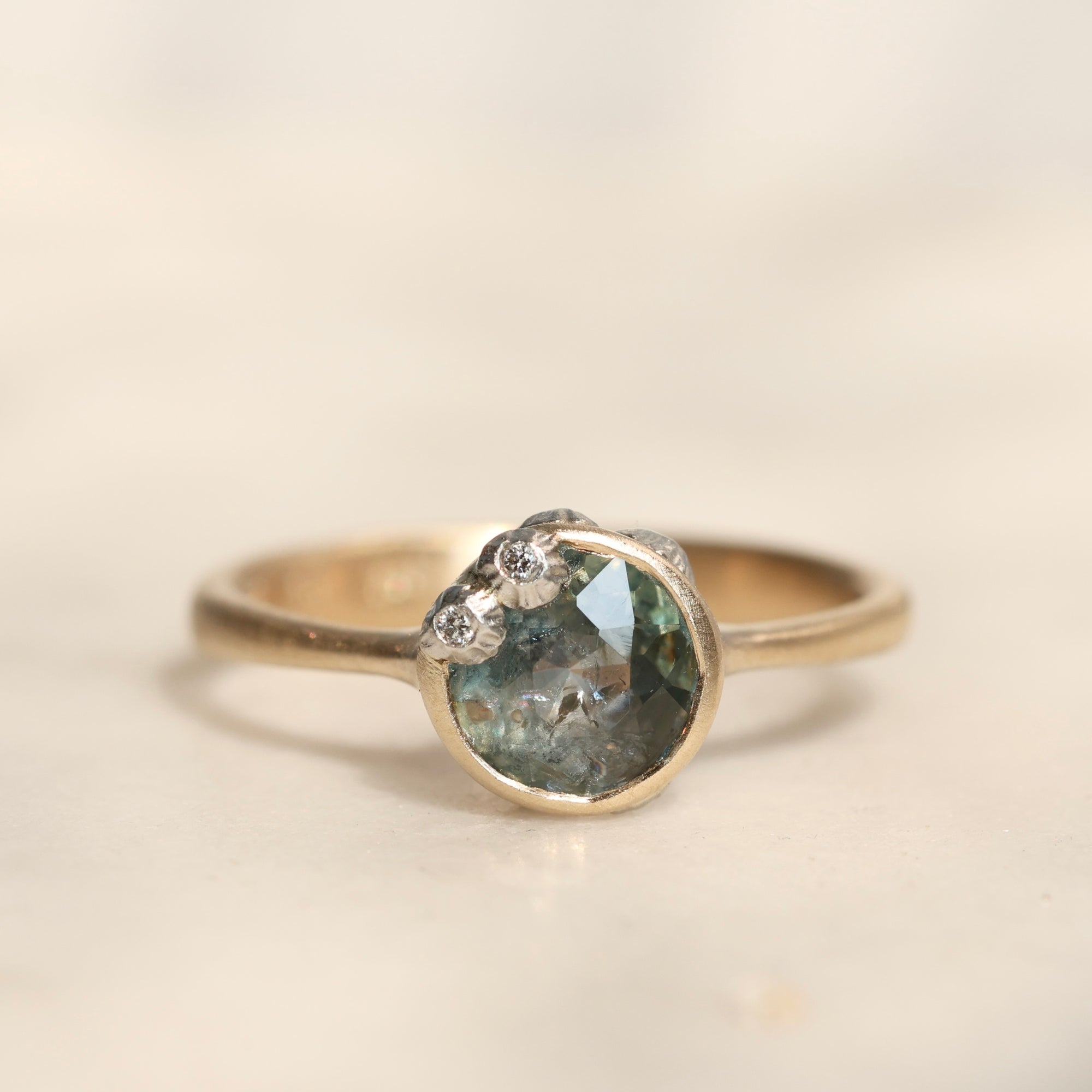 A silver ring featuring a round, faceted gray-green gemstone with small barnacles clustered along the top, set against a soft, light beige background.