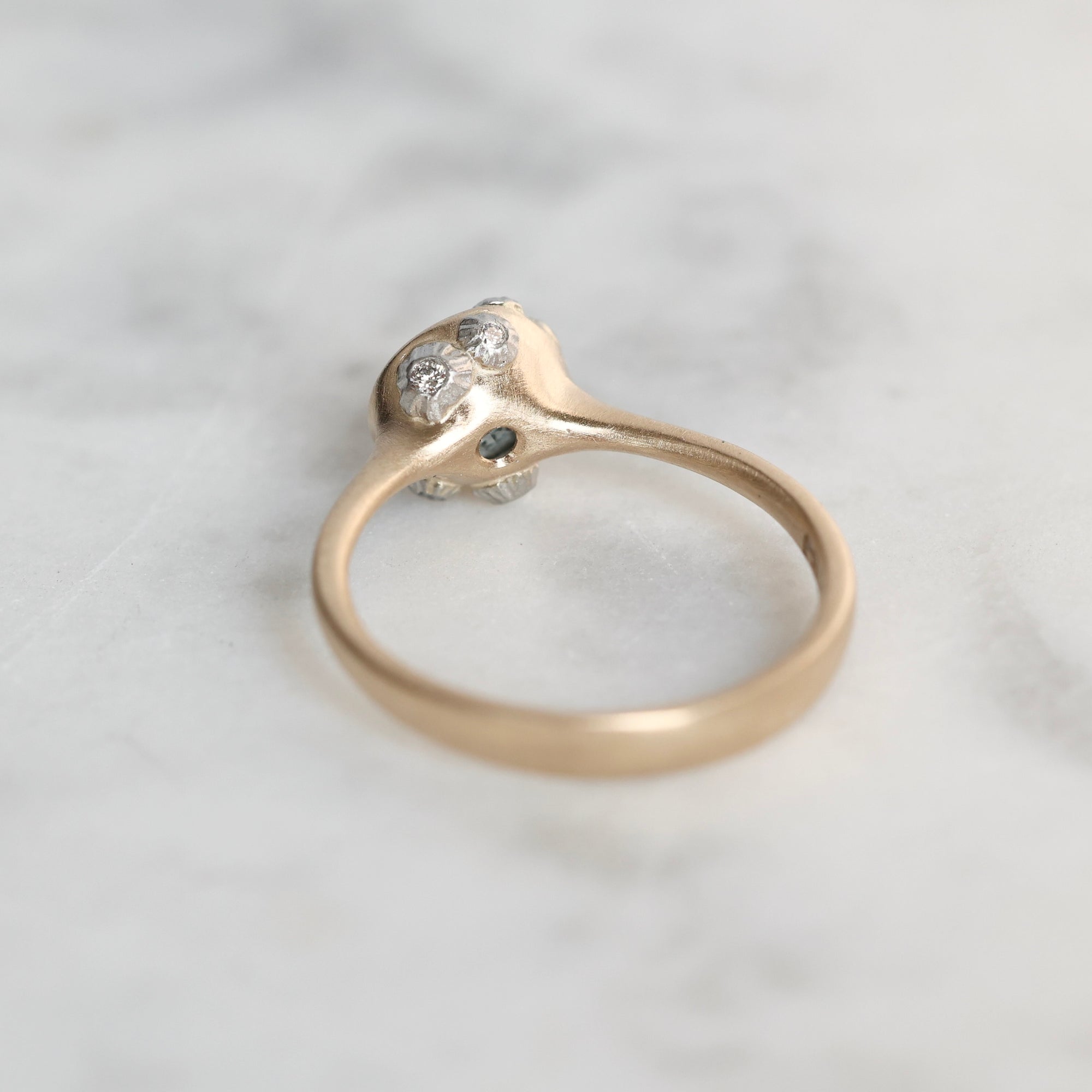 A vintage-style ring with a yellow gold band and a silver-toned setting featuring small clear gemstones, resting on a light gray marble surface.