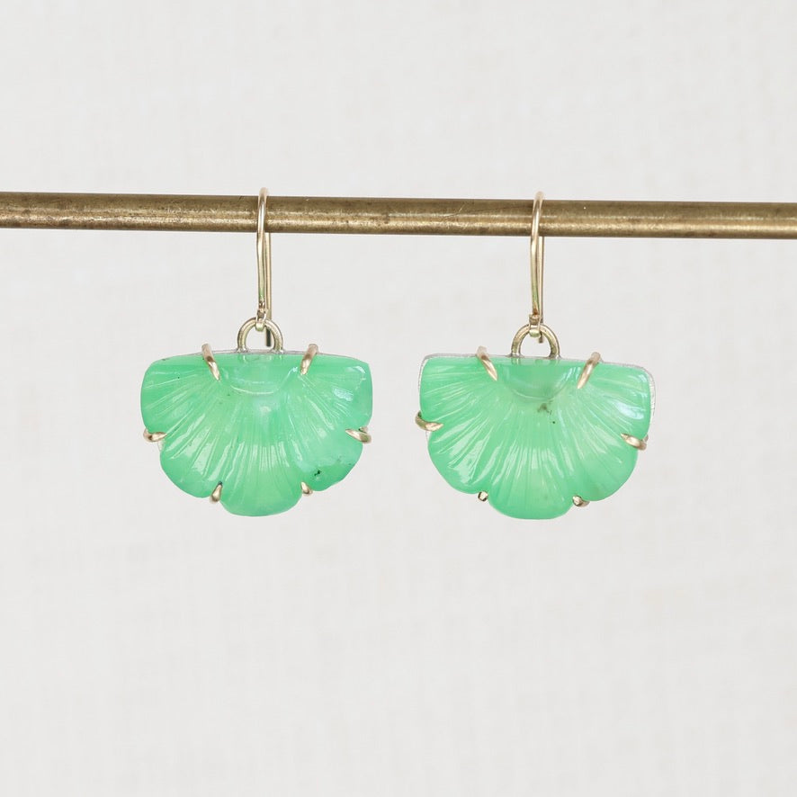 A pair of green shell-shaped drop earrings with gold hooks, hanging from a gold-colored horizontal bar against a light gray background.