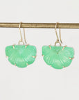 A pair of green carved stone dangle earrings with gold-toned hooks, hanging from a gold rod against a plain light background.