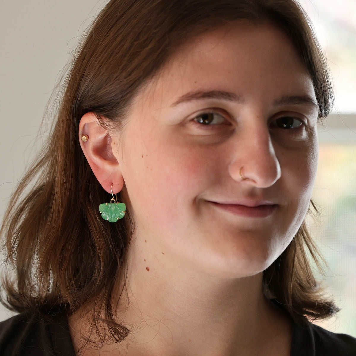 A close-up portrait of a woman wearing a small green leaf-shaped dangle earring, with her face and ear clearly visible against a softly blurred indoor background.