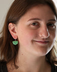 A close-up portrait of a woman wearing a small green leaf-shaped dangle earring, with her face and ear clearly visible against a softly blurred indoor background.