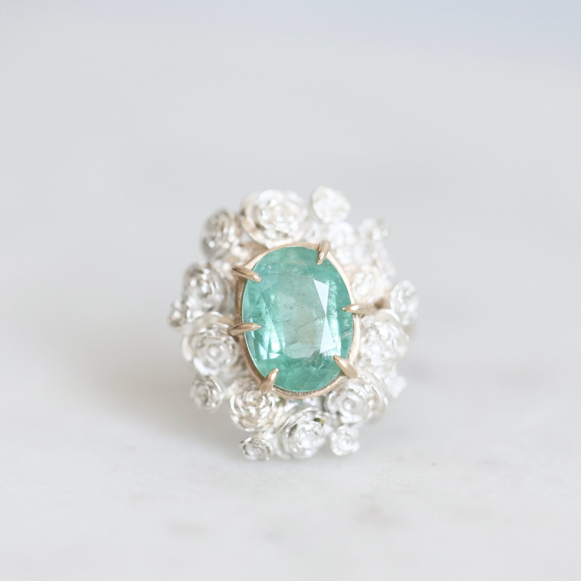 Glowing green stone held in gold prongs with a wreath of silver roses surrounding it 