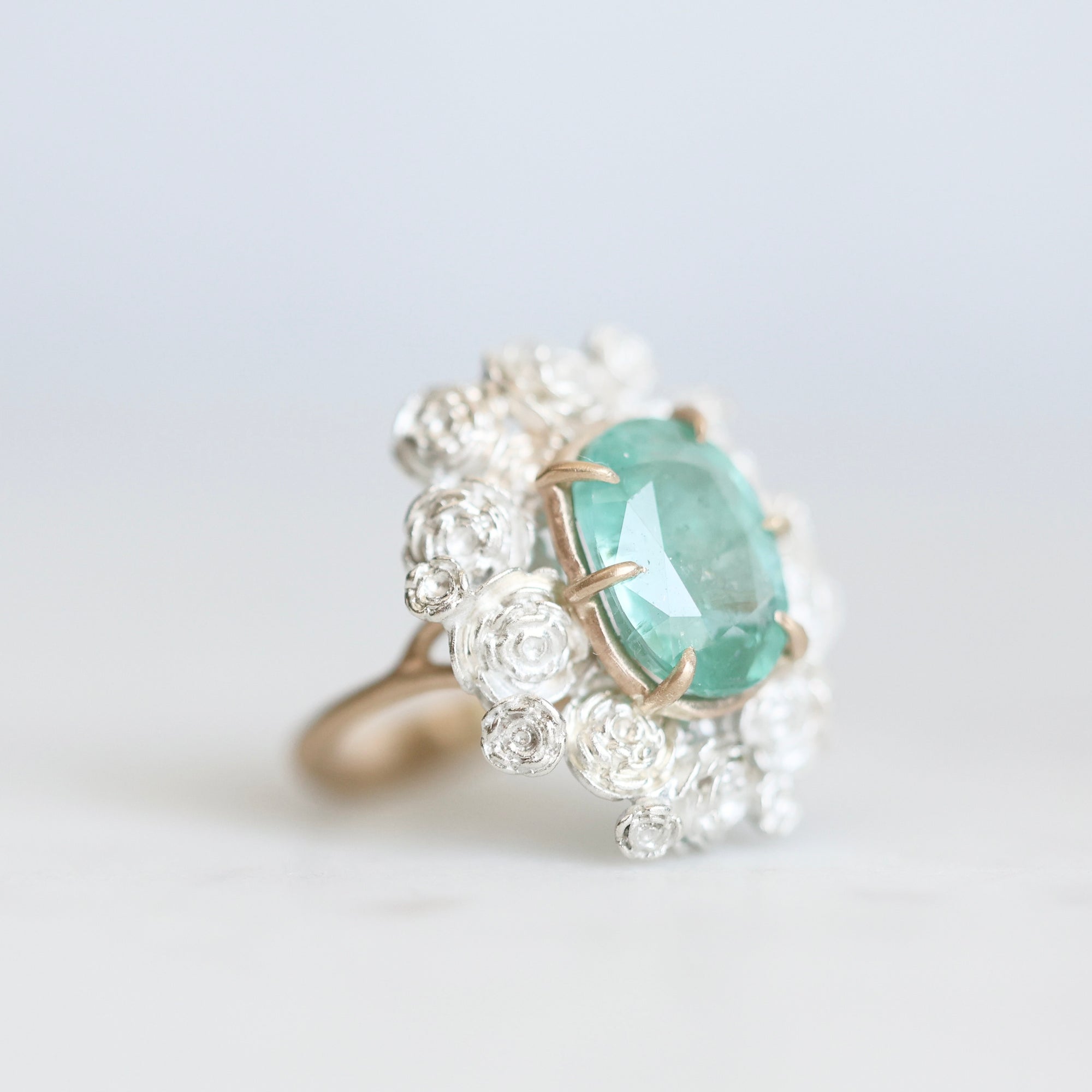 Side view of glowing green stone ring held in gold prongs with a wreath of silver roses surrounding it 