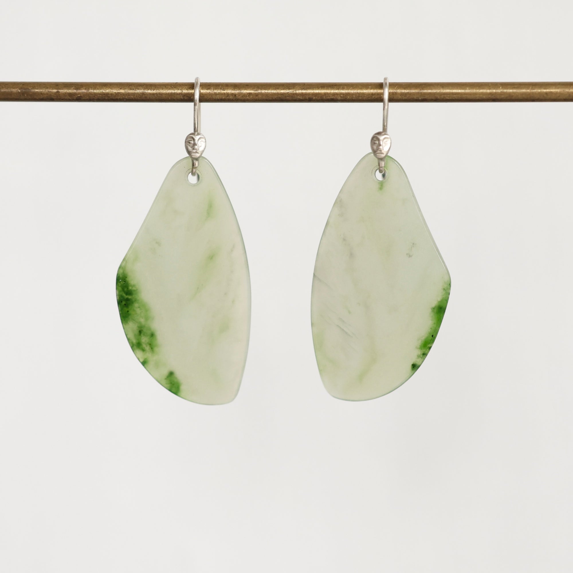 A pair of teardrop-shaped dangle earrings made from polished light green jade with natural green marbling, suspended from silver fishhook wires on a thin horizontal brass rod against a plain light gray background.