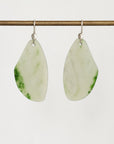 A pair of teardrop-shaped dangle earrings made from polished light green jade with natural green marbling, suspended from silver fishhook wires on a thin horizontal brass rod against a plain light gray background.