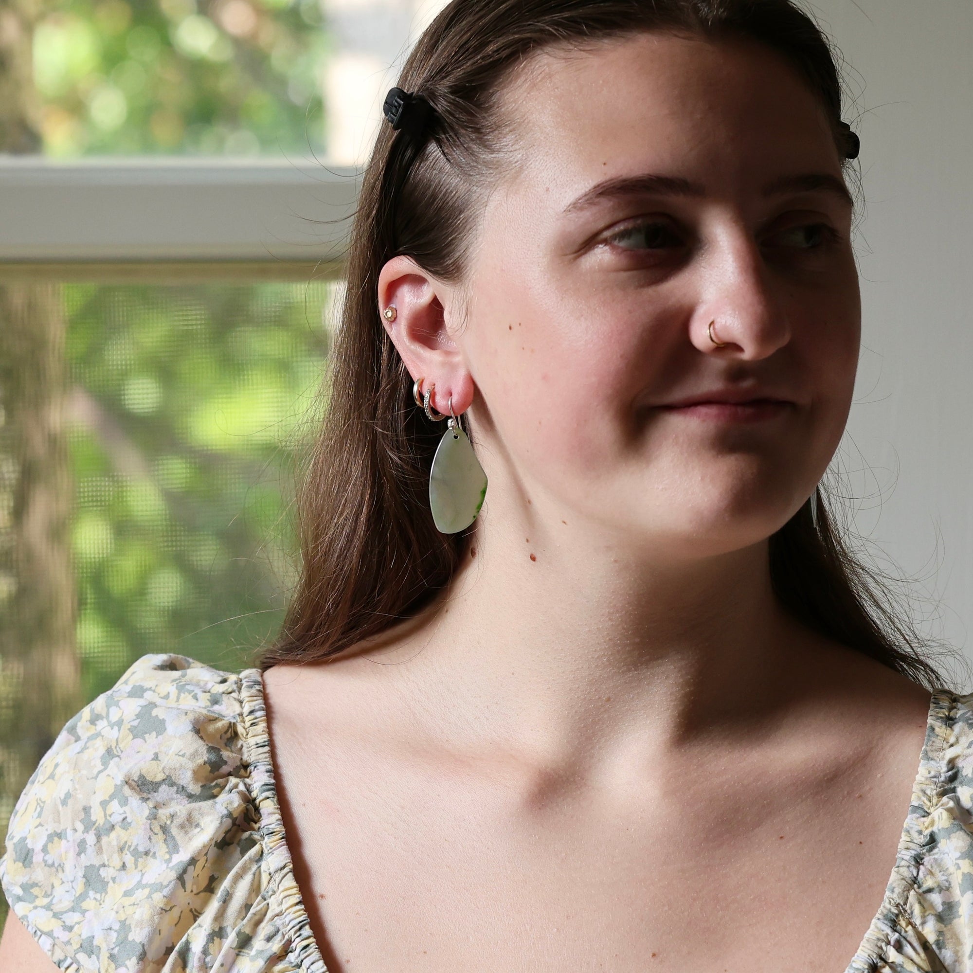 A close-up lifestyle shot of a young woman wearing a pair of handmade dangle earrings. The earrings feature a large, irregular, light green stone or resin drop on a silver-toned wire, with smaller coral-pink and silver hoop earrings stacked on her ear.