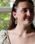 A close-up lifestyle shot of a young woman wearing a pair of handmade dangle earrings. The earrings feature a large, irregular, light green stone or resin drop on a silver-toned wire, with smaller coral-pink and silver hoop earrings stacked on her ear.