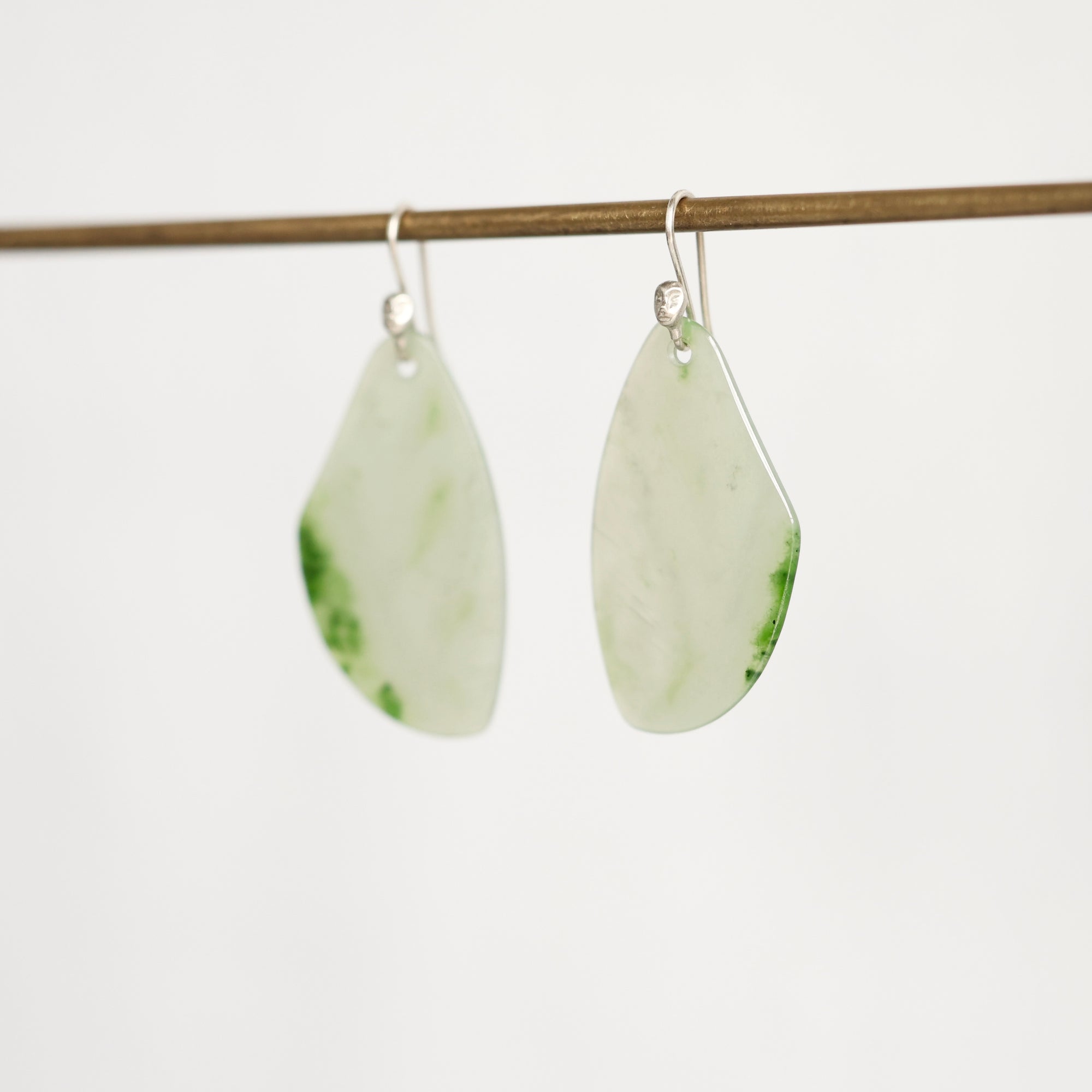A pair of teardrop-shaped earrings made from light green jade with natural green inclusions, hanging from silver fishhook ear wires on a thin wooden rod against a plain white background.
