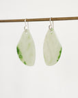 A pair of teardrop-shaped earrings made from light green jade with natural green inclusions, hanging from silver fishhook ear wires on a thin wooden rod against a plain white background.
