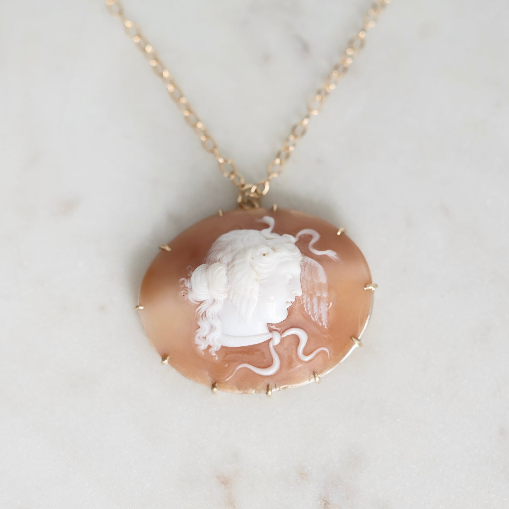 A gold-tone necklace featuring an oval carved shell pendant with a white classical bust profile on a warm peach-brown background, displayed on a light marble surface.